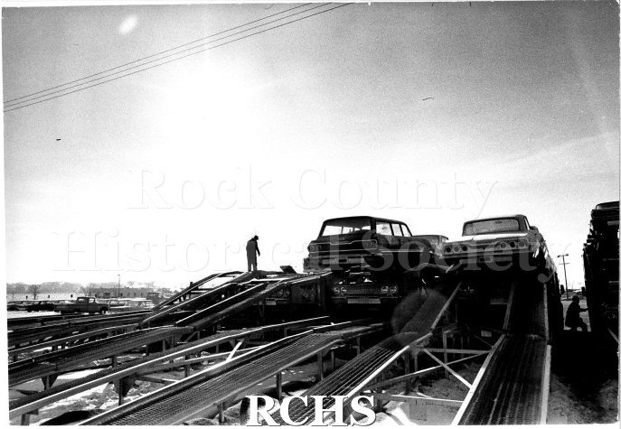 Black and white photograph of JATCO workers loading cars onto the transport trailers. Several trailers are being loaded in a row, each with two levels of cars.