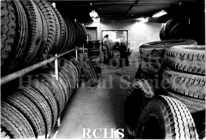 Black and white photograph of the tire bay at JATCO. Shelves of tires frame the photo. On the left is a shelf with two levels of tires and a stack of hubcabs.