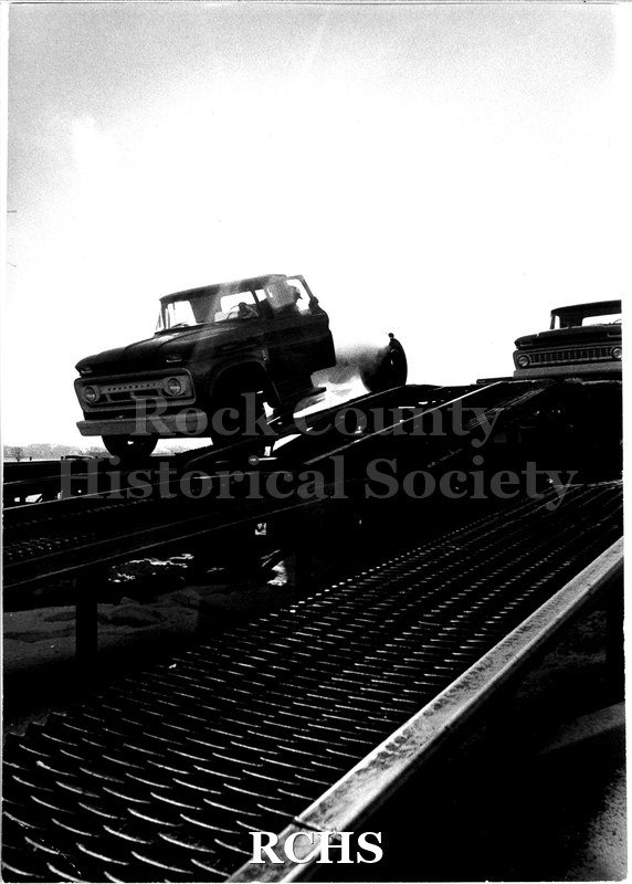 Black and white photograph of a JATCO worker backing a Chevrolet C-20 pickup truck up onto the transport trailer.