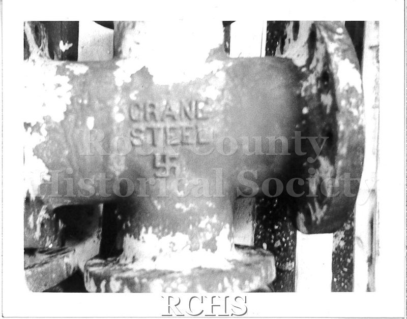 Historic image of Black and white Polaroid of a machine part from a tractor. The part is embossed with the foundry (Crane Steel) as well as a swastika underneath.