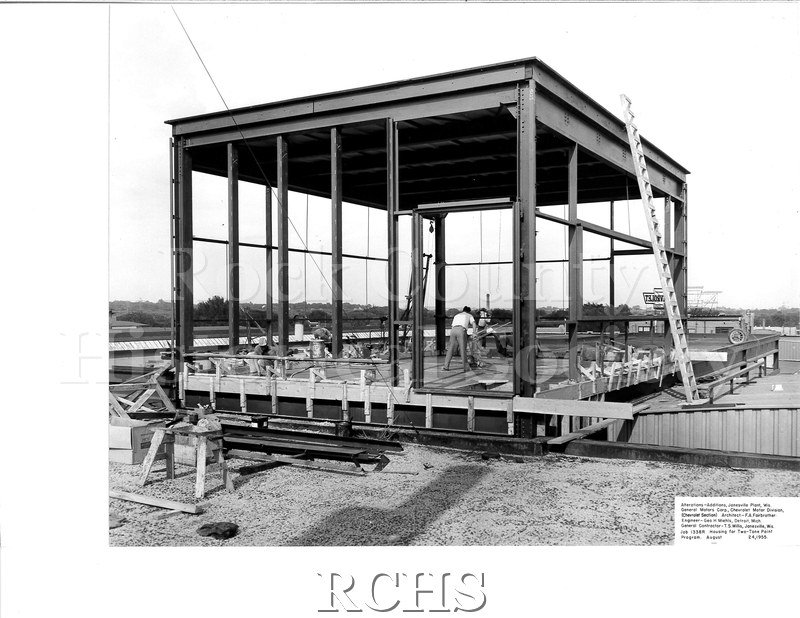 Black and white photograph of men working on a rectangular part of the roof top. There is a ladder on the right side.