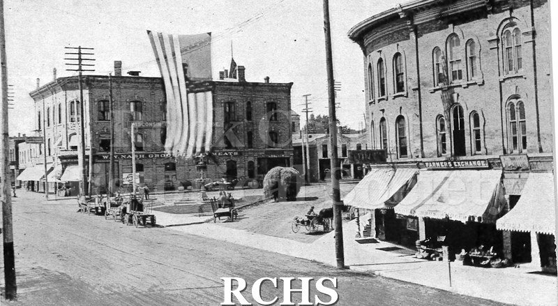 Postcard. Black and white photograph of the corner of Franklin and West Milwaukee Streets in Janesville. This corner is often referred to as the Corn Exchange.