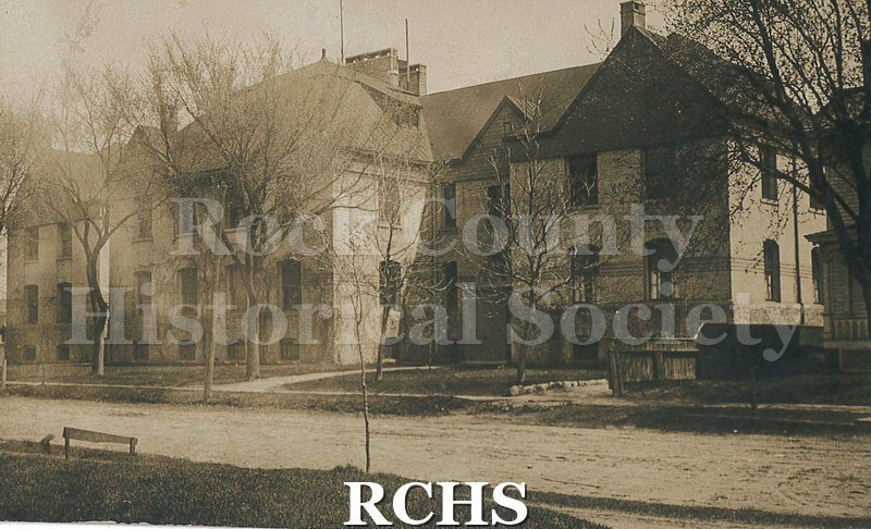 Black and white postcard of building with bare trees, grass and road in foreground. Fence on right with part of house visible. Building is 2 1/2 stories.