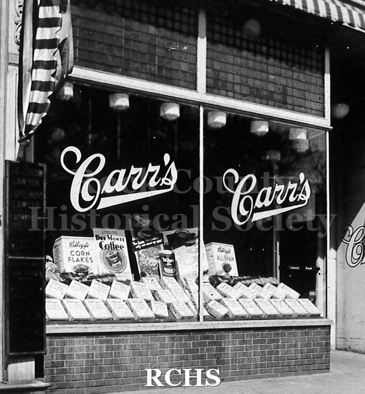 Black and white photograph of Carr's Groery Store windows. Each window has the name "Carr's" on it. A box of Kellogg's Corn Flakes and other goods are displayed.