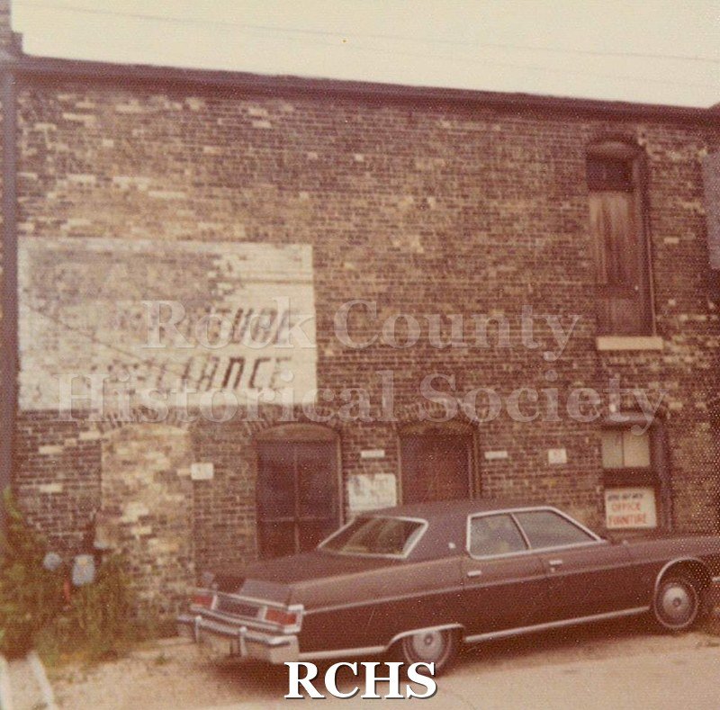 Black and white photograph of a faded sign on a brick building. There is a car parked in front of the building.