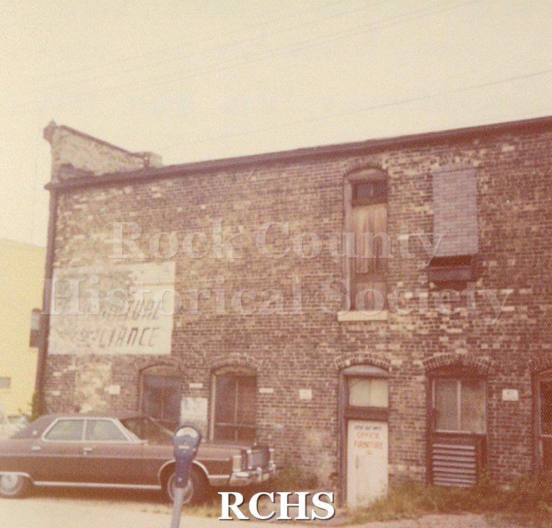 Black and white photograph of a faded sign on a brick building. There is a car parked in front of the building.