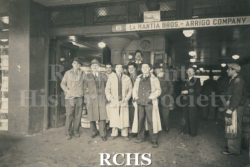 Black and white photograph of several men standing in the lobby of a building. Above their heads is a sign: "La Mantia Bros.-Arrigo Company".