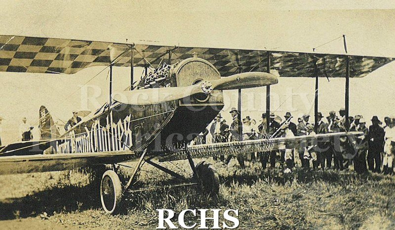 Black and white photograph of many people standing around a single engine biplane. There is a sign on the plane's left side that reads in part: "Brand Clothes".