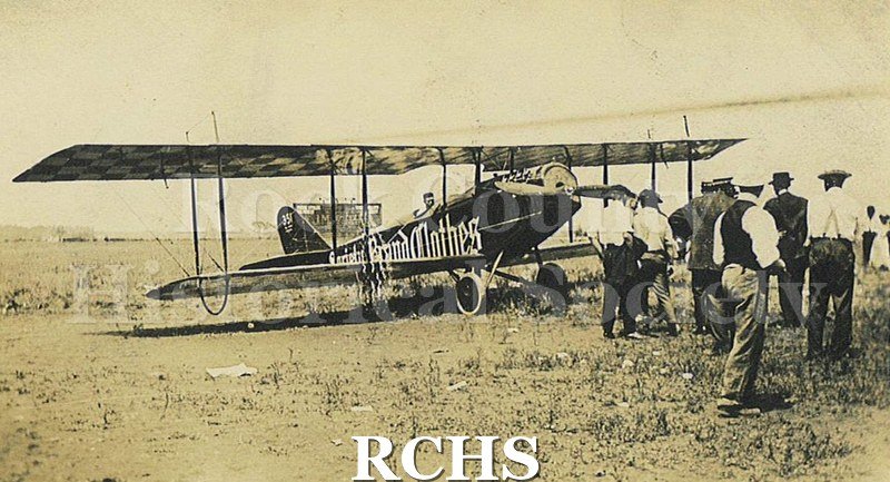 Black and white photograph of many people standing around a single engine biplane. There is a sign on the plane's left side that reads in part: "Brand Clothes".