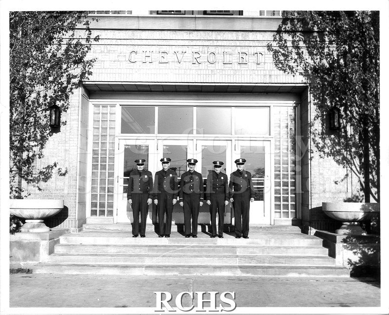 Black and white photograph of five military personell posed outside the front doors to the Chevrolet building of the General Motors plant in Janesville.