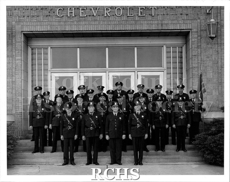 Black and white photograph of military personell posed on the steps outside of the General Motors plant in Janesville. 28 men are standing in four rows.
