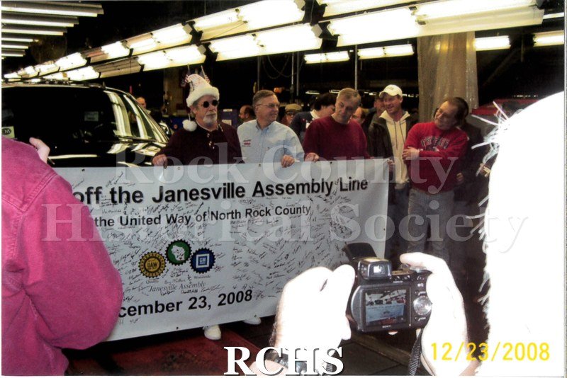 Digital color photo of employees at Janesville General Motors posing in front of the last car to come off the line at the plant. Same scene as 2022.184.5.