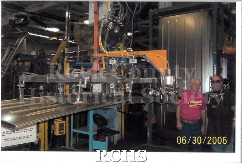 Digital color photo of two employees at Janesville General Motors posing together on the line. Same scene as 2022.184.6.
