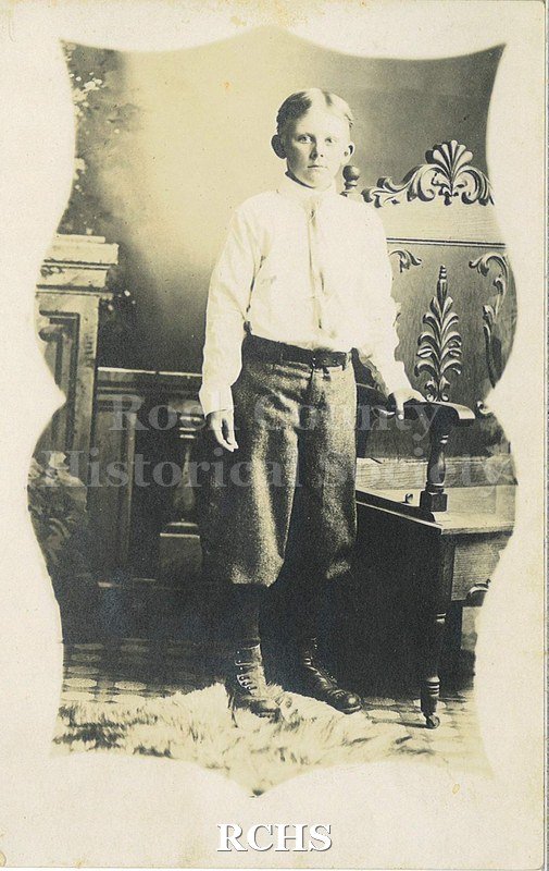 Post card with Black and white photograph of an unidentified young boy standing near a table. He is wearing a white shirt, dark pants and tie.