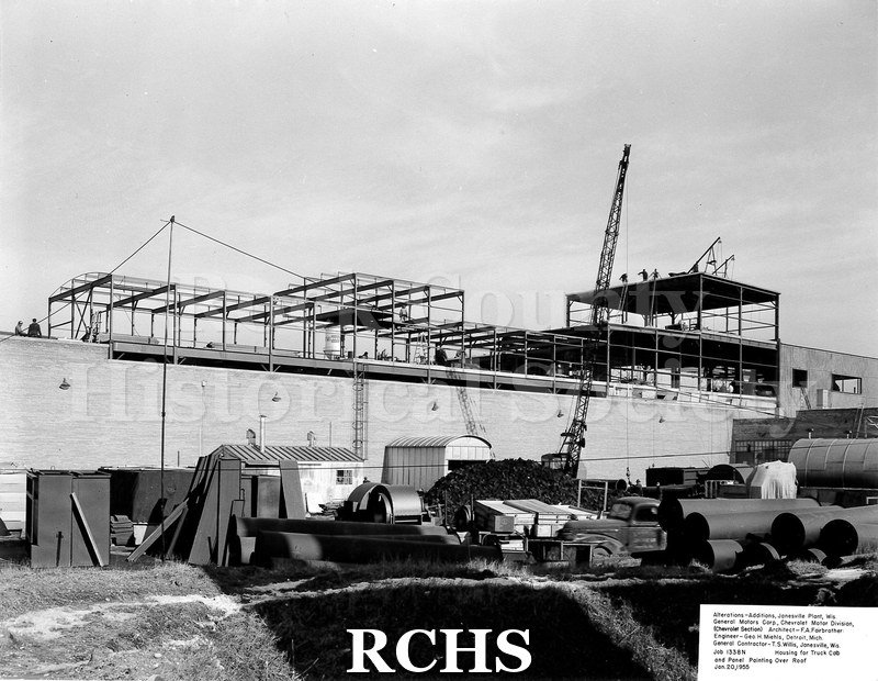 Black and white photograph of the exterior of the Chevrolet plant under construction. There is a crane in front and a truck beside large tubing.