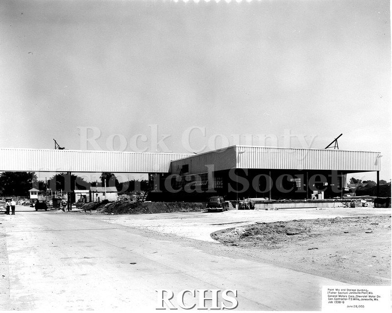 Black and white photograph of the exterior of the Chevrolet plant under construction. There are people on the road and cars parked near the building.