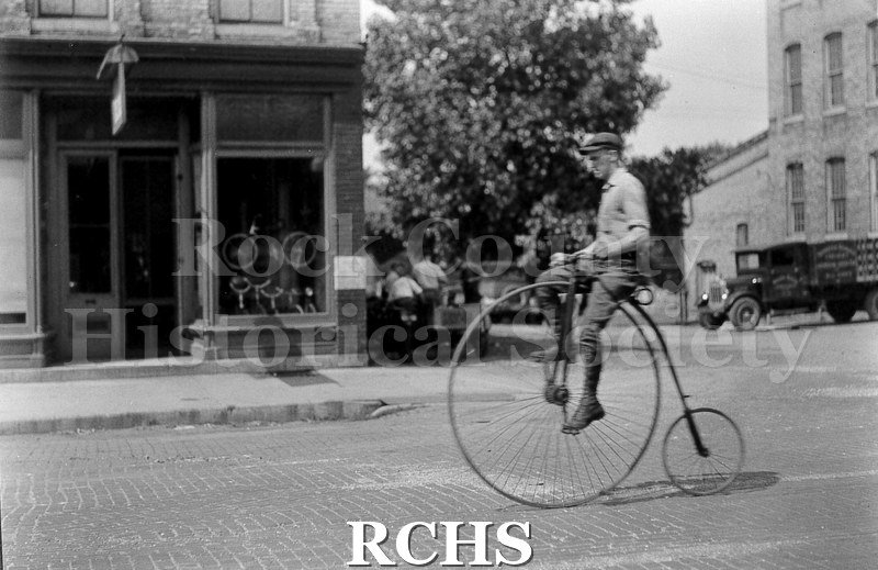 Black and white photograph of an unidentified man riding a high wheel bicycle with one large wheel in front and a small wheel in the back.