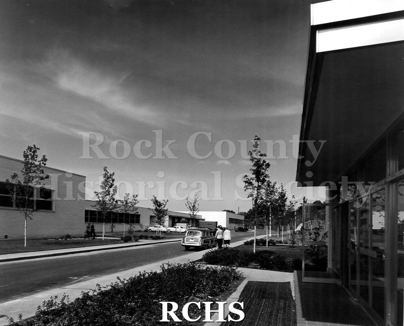 Black and white photograph of the exterior of Arrow Park Plant. There is a car on the road and two people walking on the sidewalk.