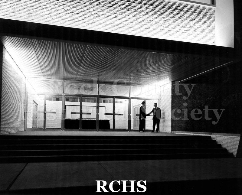 Black and white photograph of the exterior of Arrow Park Plant. There are steps going up to the door where two men are standing.