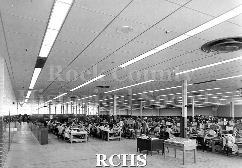Black and white photograph of the interior of the Parker Pen factory. There are employees working at machines throughout the room.