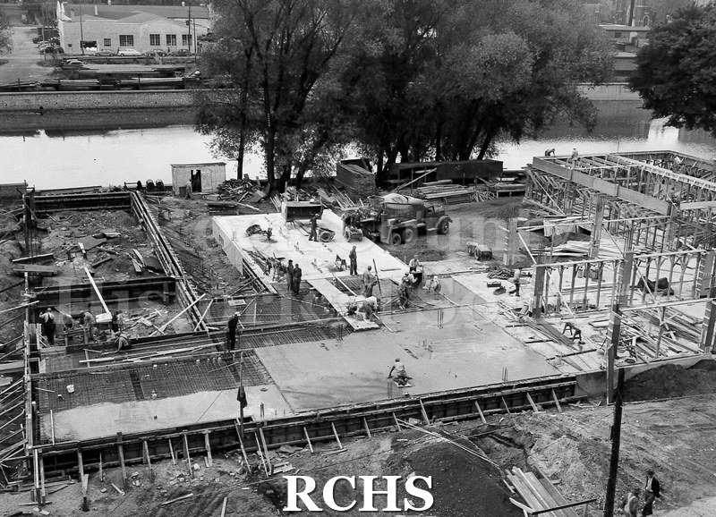 Black and white photograph of Rock County Jail under construction. The footings have been laid and men are pouring cement and spreading it out.