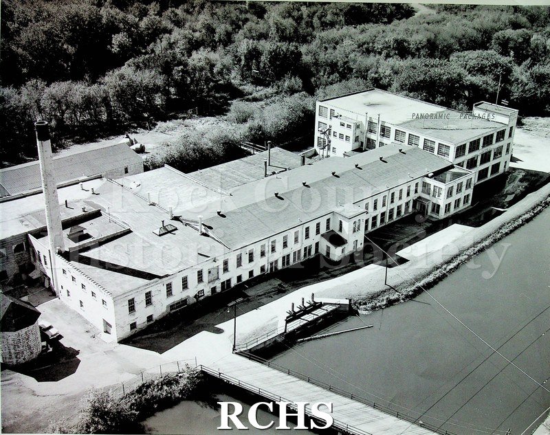 Black and white photograph of Panoramic Packages. Long white two story building with far end three stories. Smokestack of left end.