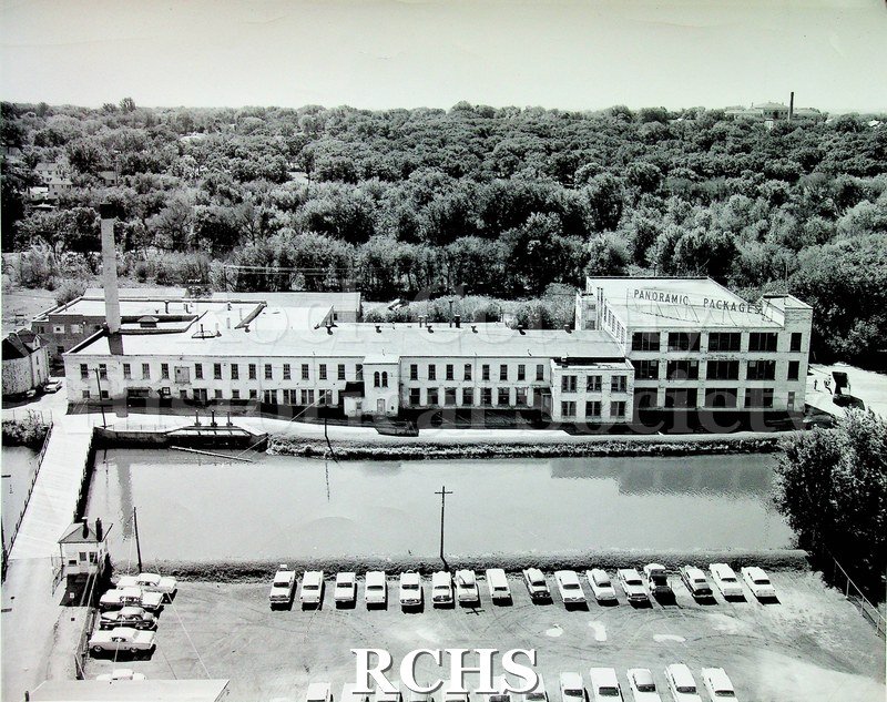 Black and white photograph of Panoramic Packages. Long white two story building with far end three stories. Smokestack of left end.