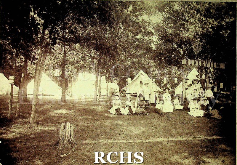 Black and white photograph of unidentified men and women at riverview campsite. 5 white tents visible, some women sitting in front, others standing in back.