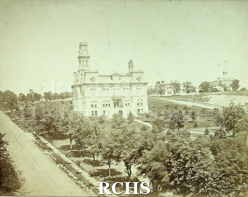 Black and white photograph of Old Janesville courthouse. Large white building 3 stories and tower facing the street. 3-4 people visible behind railing above stairs.