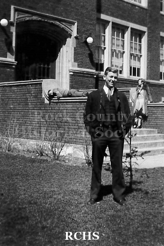 Black and white photograph of Charles N. Tallman as young man, dressed in suit, vest and tie. Standing in front of brick building with steps.