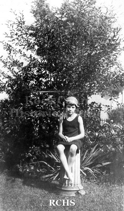 Black and white photograph of Elizabeth C. Tallman as young girl dressed in bathing suit with cap, seated on white pedastal in garden.