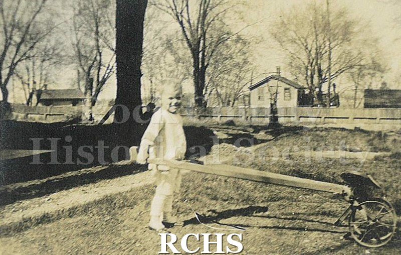 Black and white photograph of Charles Norton Tallman dressed in white holding wood pull to child's wheeled seat. Houses and trees in bakground.