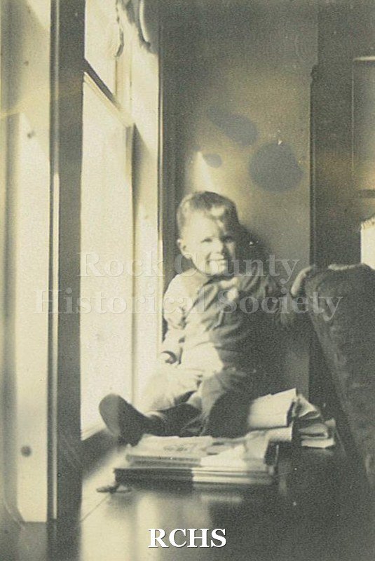 Black and white photograph of Charles Norton Tallman dressed in long sleeved shirt and pants seated on window seat with windows to his right.
