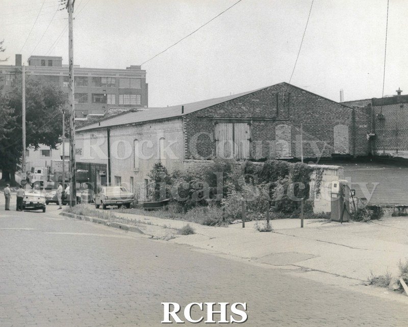 Black and white photograph of the Gazette building. Shows two brick buildings. There is a gas pump standing alone on the right side.