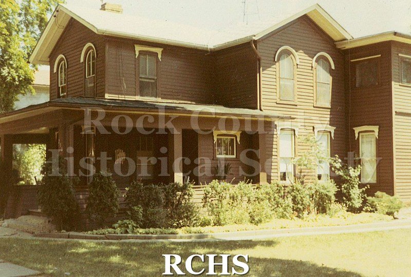 Colored photograph of an unidentified two story house on Madison St., Janesville, WI. Shows wrap-around porch with shrubs in front.
