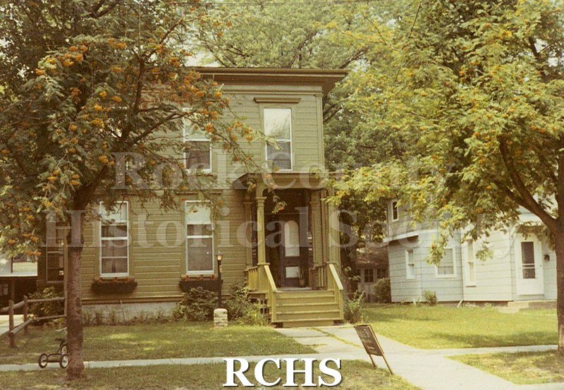 Colored photograph of an unidentified two story square brown house on Madison St., Janesville, WI. Shows small porch with trees in front.