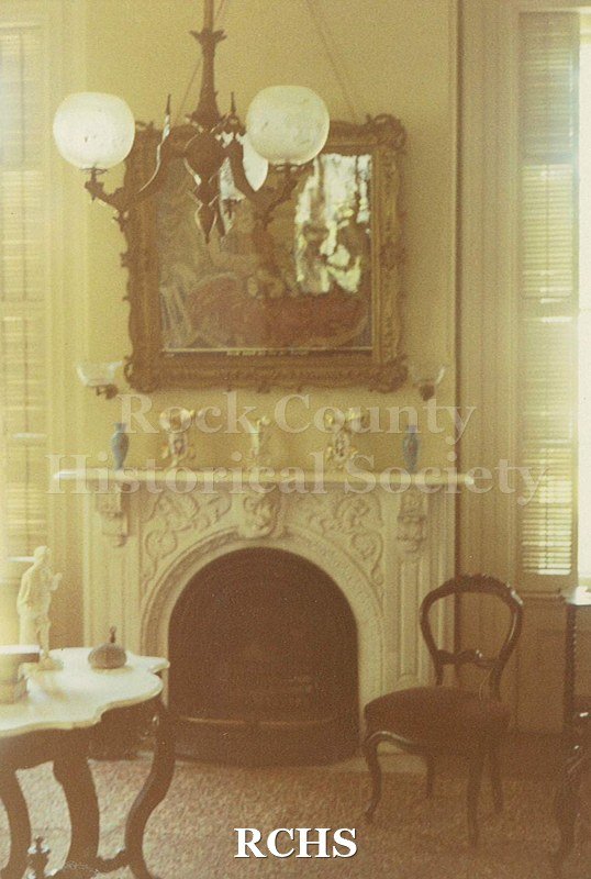 Colored photograph of the inside front room of the Tallman House. Shows the fireplace with picture over it; gas lights and table with marble top.