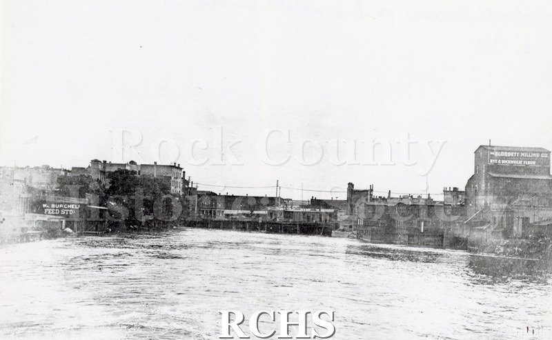 Black and white photograph of the Rock River looking south from Fourth Avenue bridge. Burchell Feed Store and Blodgett Milling Company are visible.