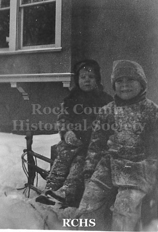 Black and white photograph of Elizabeth & Charles N. Tallman seated on sled in front of the bay window of the Charles Tallman House.
