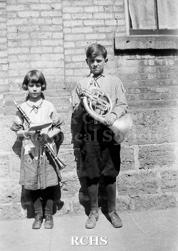 Black and white photograph of Elizabeth & Charles Tallman standing in front of brick and block sided building with window with curtain, both holding musical.