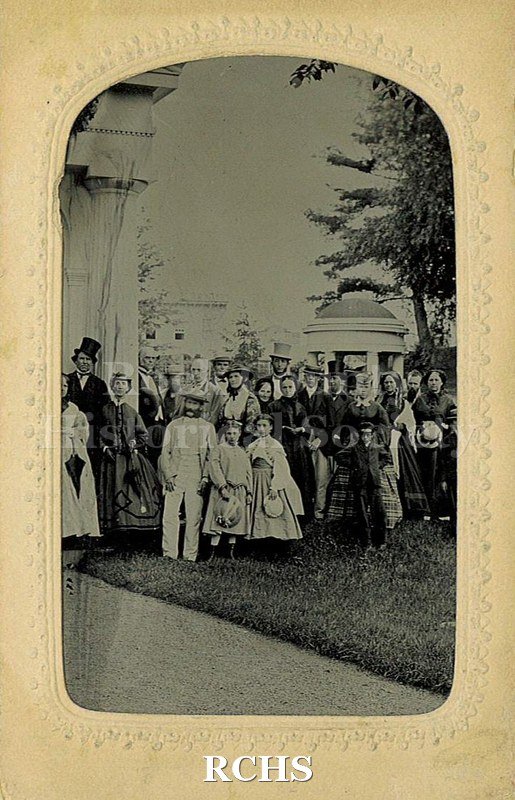 Black and white photograph of a group of men, women and children on grass in front of buildings with pavilion behind. Men in suits and hats.