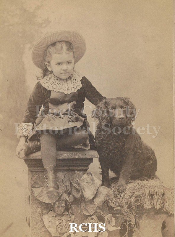 Black and white photograph of May Felton as a young girl seated on column with black down to her left.