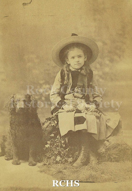 Black and white photograph of May Felton as a young girl seated on "rock" with flowers and moss, "grass". She is wearing a broad brimmed straw hat.