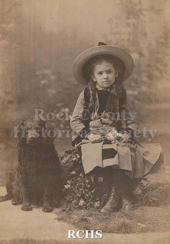 Black and white photograph of May Felton is the young girl seated on "rock" with flowers and moss, "grass". She is wearing a broad brimmed straw hat.
