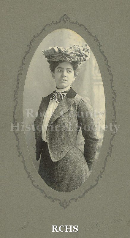 Black and white photograph of May Felton in 3/4 left profile body and full on head. Eyes looking to her left, hat perched on top of head with large straw flowers.