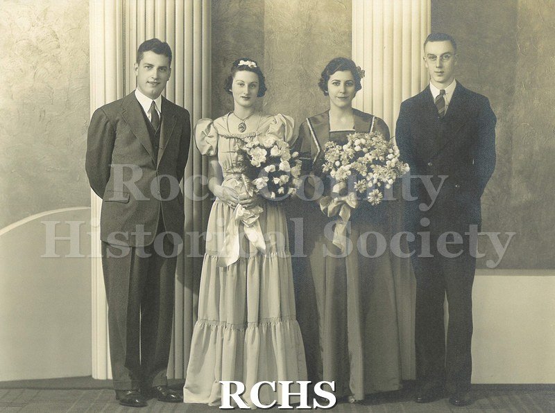 Black and white photograph of a Wedding party. Groom on left in dark suit, vest, tie with white shirt, standing with arms behind his back.