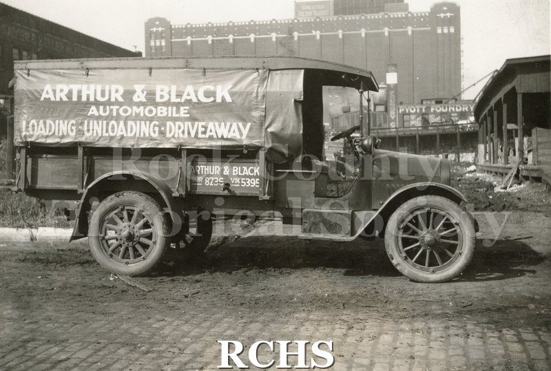 Black and white photograph of an "Arthur & Black Automobile" truck advertising loading, unloading, and drive-away. Truck is parked in a large city.