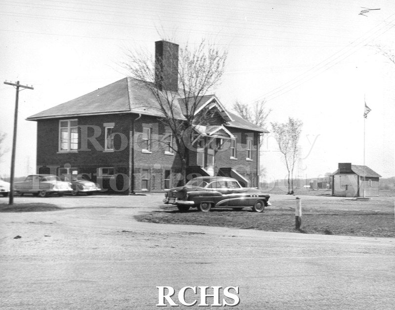 Black and white photograph of a street view of what is possibly the Janesville Consolidated School. Four cars are parked by the school.