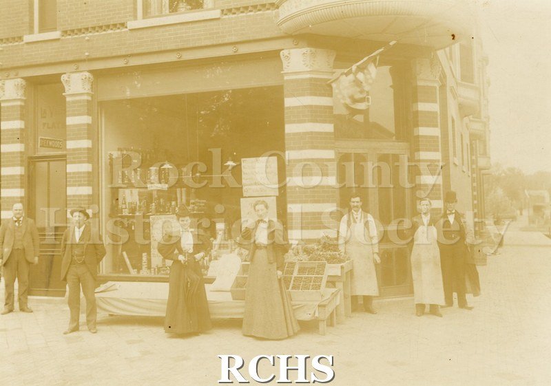 Tinted photo of 50 S. Main St., Janesville, WI. Shows a store with seven people standing in front of it. There is fruit in boxes on display in front of the store.