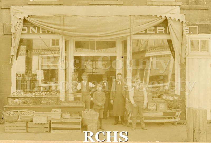 Tinted photo of Jones' Grocery Store with four men and a boy standing in front of it. There is merchandise on tables and crates in front of the store. .